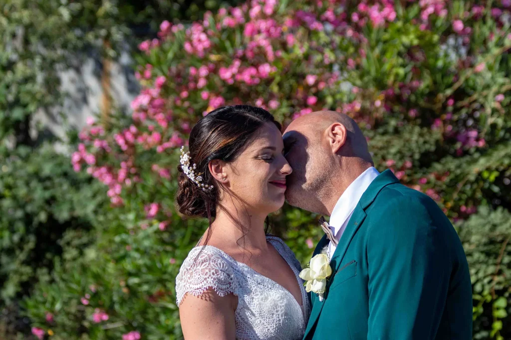 Wedding couple sharing a tender kiss during a photoshoot in Provence, with the bride in a white lace dress and the groom in a green suit, surrounded by greenery and pink flowers