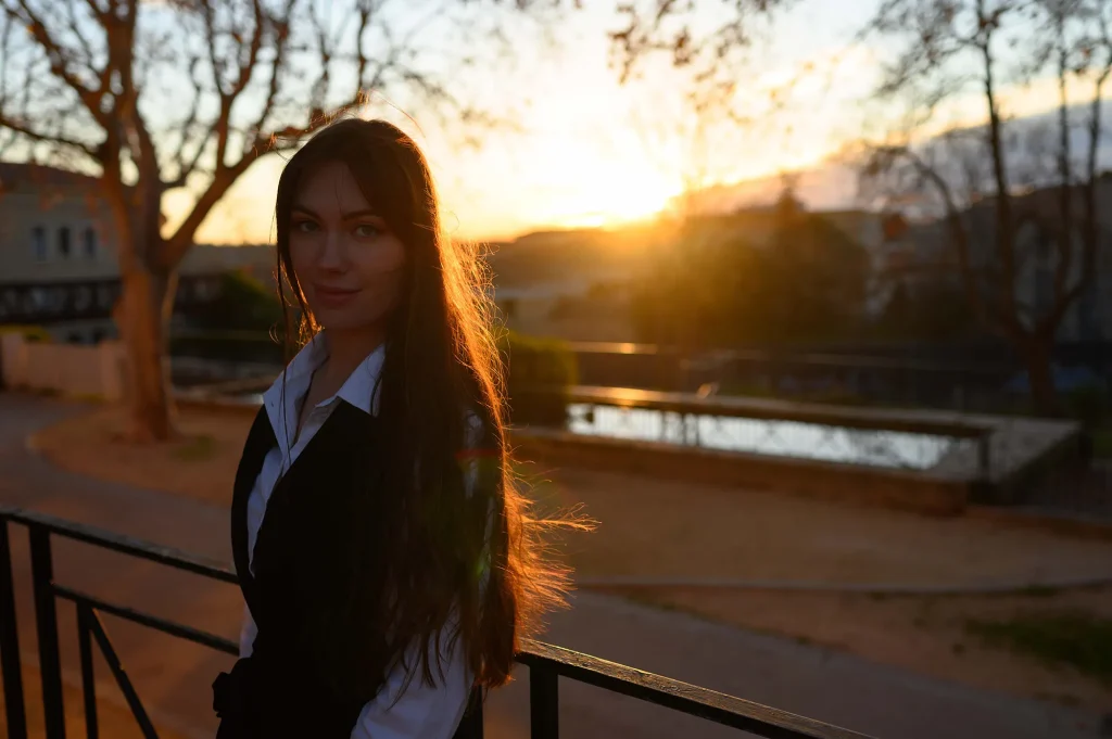 Elegant woman in a black dress and white blouse, with long hair, posing at Parc Jourdan in Aix-en-Provence at sunset in winter, with a pond in the background