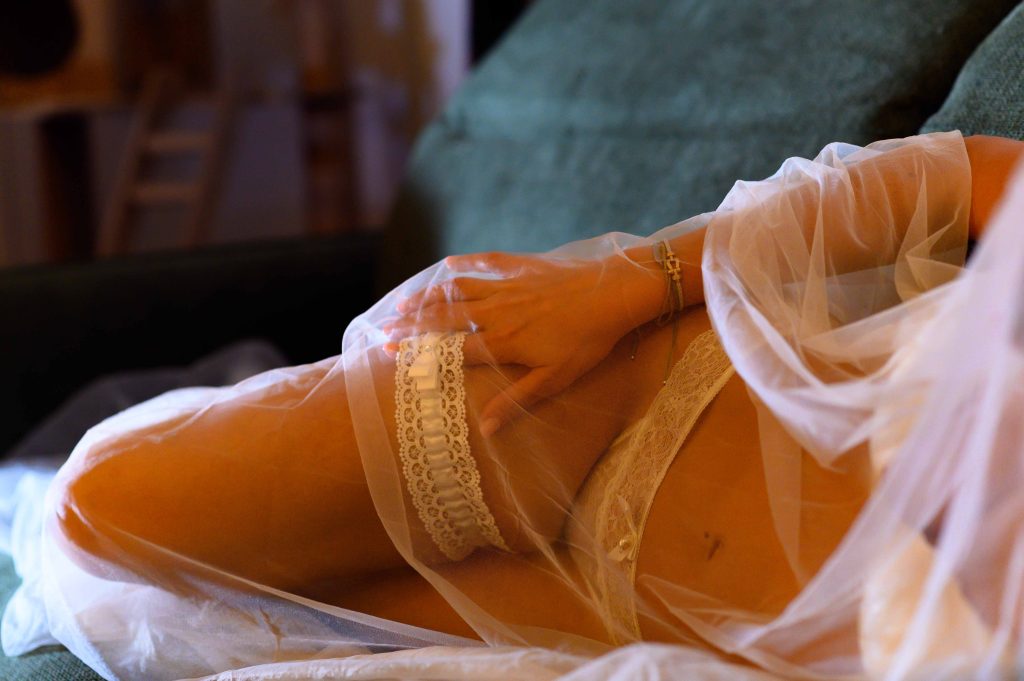 Future mariée allongée sous un voile avec la main posée sur la jambe pendant une séance bridal boudoir