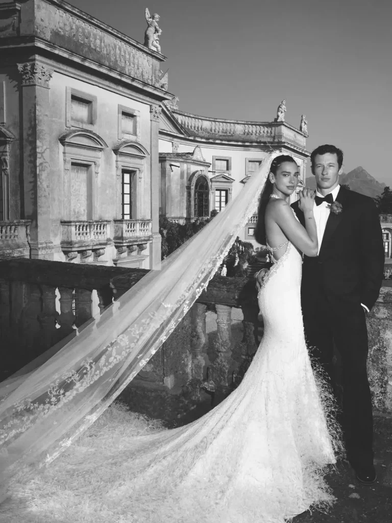 Dua Lipa in a long train wedding dress and Callum Turner in a tailored suit, posing in front of a historic villa in Palermo, Sicily