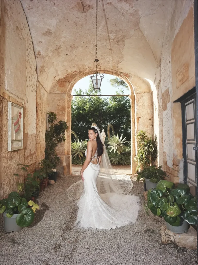 Dua Lipa in a stunning backless wedding dress walking through a rustic courtyard with lush greenery in Palermo