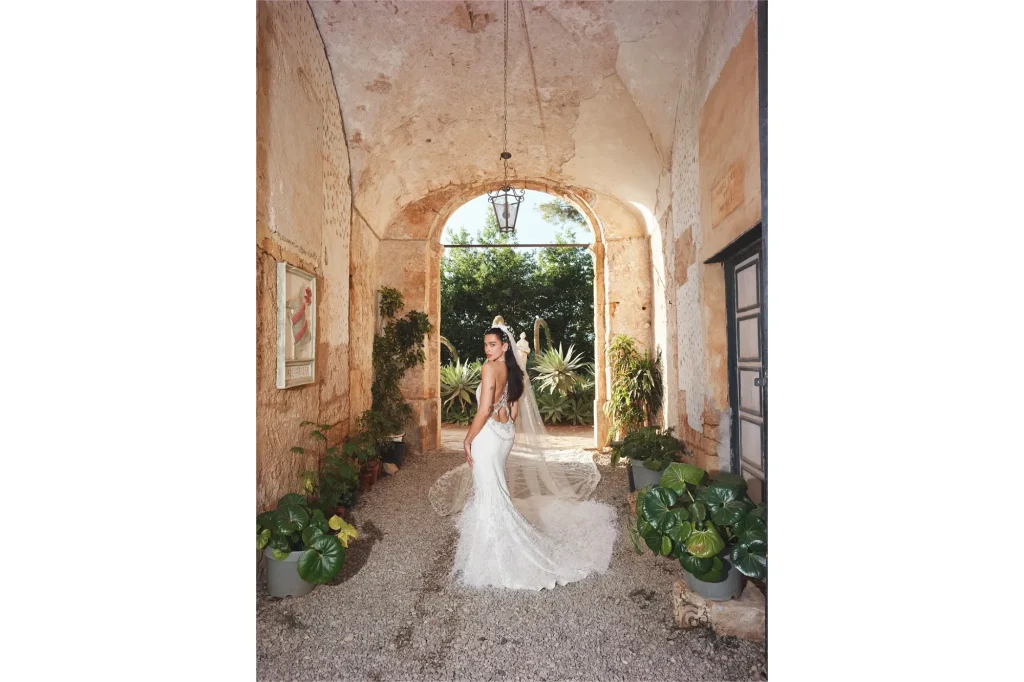 Dua Lipa in a stunning backless wedding dress walking through a rustic courtyard with lush greenery in Palermo