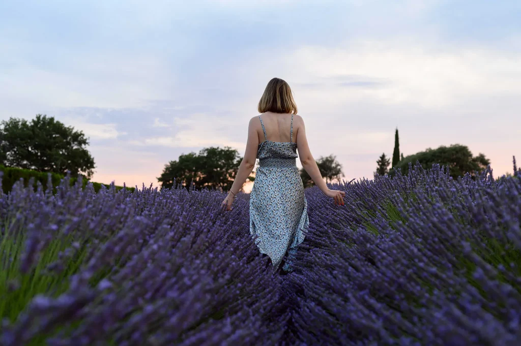 Femme en robe longue à motifs floraux marchant dans un champ de lavande en Provence au coucher de soleil, vue de dos avec les bras légèrement écartés, ambiance sereine et lumière dorée