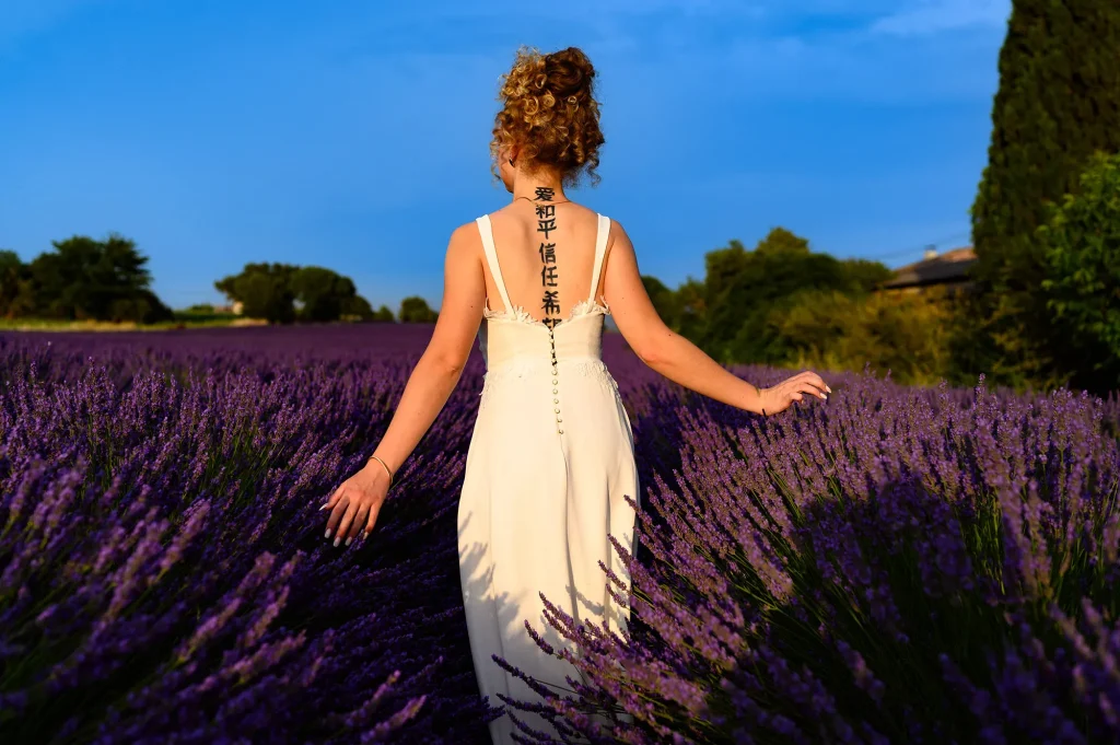 Mariée en robe blanche dos nu avec tatouage, marchant dans un champ de lavande en Provence, vue de dos avec les bras écartés, lumière naturelle et ciel bleu