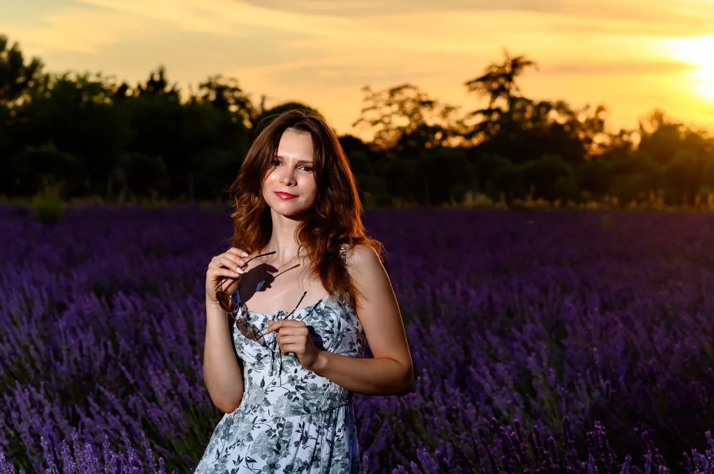 Young woman in a floral dress standing in a lavender field in Provence at sunset, with golden light and a warm orange sky